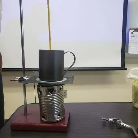 A tin can cookstove is sitting on a ring stand. A ring clamp is attached to the ring stand and wire mesh is sitting on the ring clamp. The ring clamp and wire mesh are sitting directly on top of the cookstove.  A steel mug is sitting on top of the wire mesh.