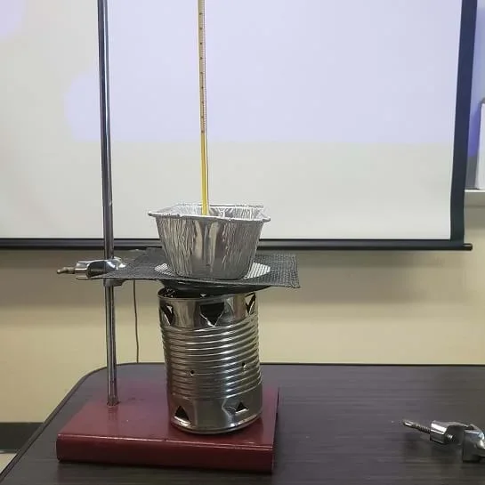A tin can cookstove is sitting on a ring stand.  A ring clamp is attached to the ring stand and wire mesh is sitting on the ring clamp. The ring clamp and wire mesh are sitting directly on top of the cookstove.  An aluminum loaf pan is sitting on top of the wire mesh.