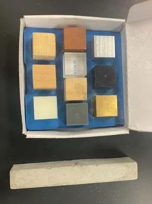 The image shows the strength test station. The buckets being used are holding up the concrete sample so that weights can be hung suspended around the sample to test how much weight the sample can hold up. A string is tied to the middle of the sample where a divot is made to hold the string in place and the weights are tied to the other end being held suspended by the concrete sample. The weights are increased until the concrete sample breaks. The image shows the sample holding up a 200g weight to the left and a 5 lb. weight on the right. Maximum weight added is 10lbs.