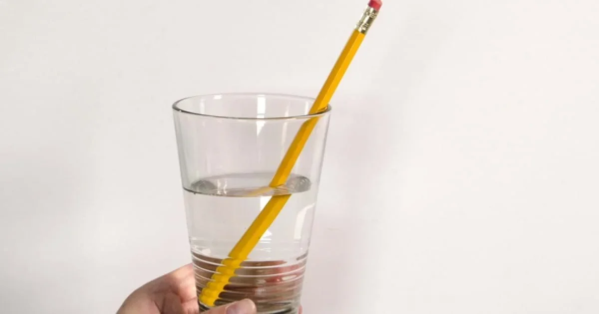 A photo of a pencil sitting upright inside a clear glass half filled with water. The pencil appears broken at the water/air interface.