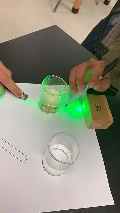 Students perform the experiment. One student shines a laser through a glass of water, and another uses a pen to draw the path of the light on the placemat.
