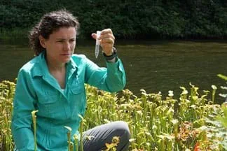 Photograph shows a young woman outdoors by a stream examining a vial of clear liquid.
