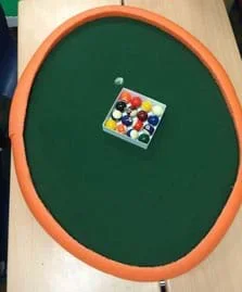 A photograph shows a view from above an elliptical-shaped pool table with a set of pool balls on a green surface material with a curved orange foam “curb” border around its perimeter.