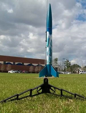 A photograph shows a model blue rocket ready to launch. The rocket sits on a grassy field next to a building.