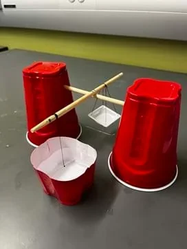 A photo showing the balance for the Method 1 activity. It is a balance beam made from plastic cups, wooden dowels, thread, needle, and weigh basket.