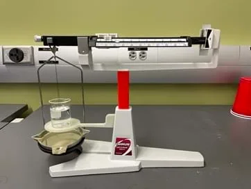 A photo showing the balance for the Method 2 activity. It is a modified triple-beam balance with a suspended button and a beaker.