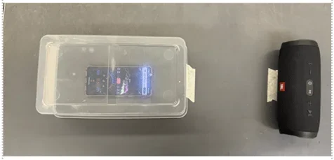 A smartphone displaying a decibel app inside a clear plastic container, next to a black JBL Bluetooth speaker, set up for a sound experiment.