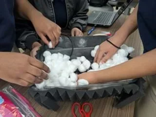 A photo of three people's hands adding cotton balls to a container lined with black sound-dampening foam.