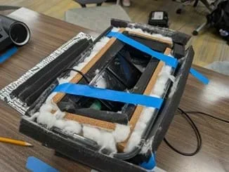 A photo of an open box filled with layers of sound-dampening materials, including foam, cotton, and cork, with blue tape holding them in place.