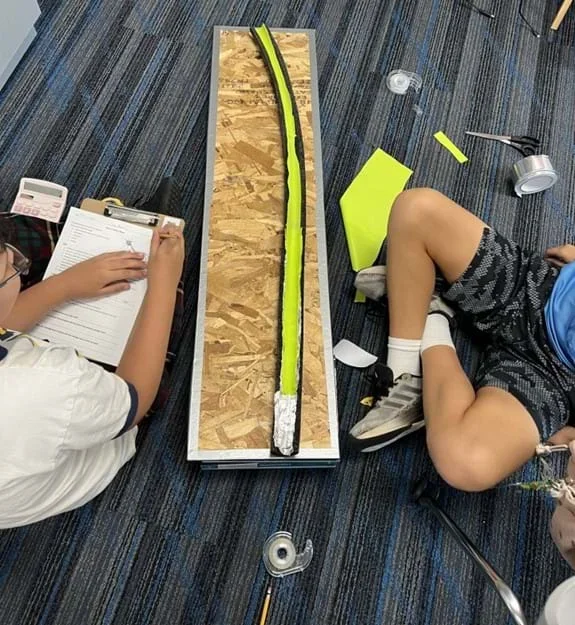 A photo showing two students seated next to a foam tube covered in rubbery material. One student is writing on a data sheet.