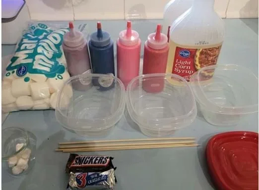 A photo showing materials for a candy-dipping activity. On the left are a bag of large marshmallows and four squeeze bottles containing different colored liquids. Behind the containers is a bottle of light corn syrup. In front are three empty clear plastic containers, two wooden skewers, a small pile of cotton or paper pieces, and a fun-size Snickers candy bar.
