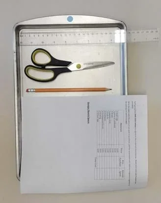 A photo of a metal tray holding scissors, a ruler, a piece of paper, and a pencil.