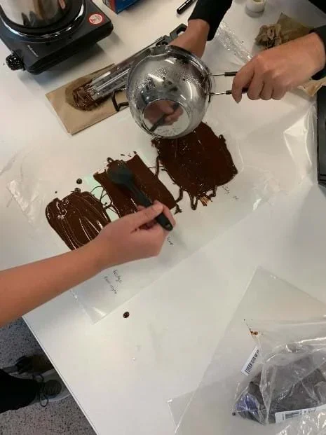 Students create holographic chocolate. They pour melted chocolate onto a piece of wax paper and the diffraction grating. The diffraction grating has been taped to the wax paper so it is still and flat while the chocolate is poured over it.