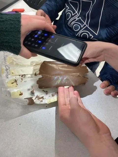 Students shine the flashlight on their phone at the chocolate. A rainbow hologram is visible on the chocolate.