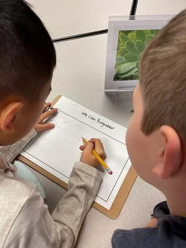 Two students stand over a worksheet on a table. One student is holding a pencil above the paper.