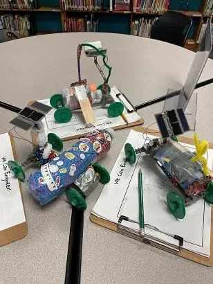 Three solar rover designs made of recycled materials on top of frames with four wheels sit on the table. Each has a small solar panel attached to the back that extends above the rover. The recycled materials for each include a tape- and sticker-covered water bottle, a small plastic bottle with popsicle sticks taped to it, and a tape-covered water bottle with a pipe cleaner attached to it.