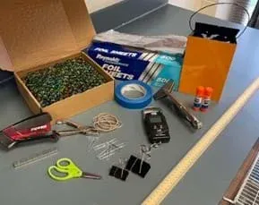 A photo of a box of foil sheets, stapler, staples, a pair of scissors, blue painter’s tape, a box of marbles, orange bucket, sheet metal vise grips, two glue sticks, a meter stick, two large binder clips, a hole punch, rubber bands, and a hanging scale.