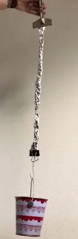 The teacher is holding sheet metal vise grips. The sheet metal vise grips are attached to the aluminum rope. At the bottom of the aluminum rope is a binder clip, S hook, and a bucket. Everything is hanging from the sheet metal vise grips.