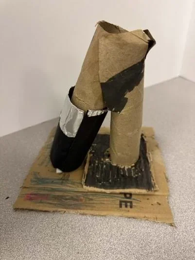 A photo of a DIY microscope model constructed primarily from cardboard materials is displayed on a flat surface. The base consists of a square piece of corrugated cardboard that has been drawn on with various colors, including red, green, and black. Two cardboard tubes are attached vertically to the base—one taller and slightly bent at the top, possibly representing the microscope's eyepiece, and another shorter tube wrapped in black paper and silver duct tape, likely symbolizing the objective lens. Black paint or marker has been used to add details on the tubes and base, giving it a more realistic microscope appearance.