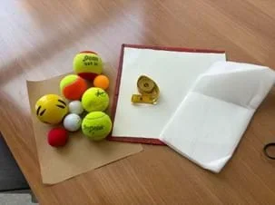 A photo showing several types of balls (tennis balls, ping pong balls, a stress ball, and a foam ball) and a rolled-up tape measure. The items are sitting on a few different surfaces that are fanned out: a sheet of sandpaper, a small dry erase board, and sheet of foam.