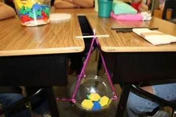 A photo showing a plastic cup suspended between two classroom desks using pink string and a wooden stick. The stick rests across the gap between the desks, with the cup hanging below it. Inside the bowl are several counters.