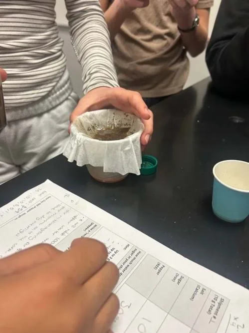 A photo showing a group of students filtering their dissolved salt and sugar aqueous solution through a coffee filter. This demonstrates the “REE purification step” in mining.