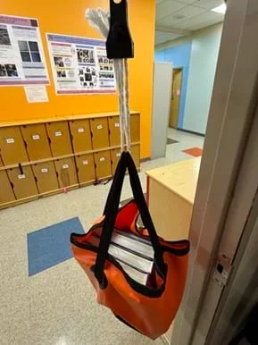 A photo of a classroom scene showing a hanging test of paper-strap prototypes. A black spring-scale-style clip at the top suspends three twisted white paper strands that are tied to the black fabric handles of an orange tote bag. The bag is filled with several thick hardcover books and hangs in a doorway above linoleum floor tiles. In the background are wooden cubbies, colorful wall posters, and a light-blue hallway.