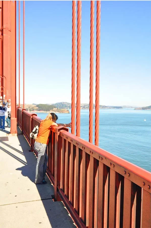 A photograph of a close-up view of the steel suspender ropes of the Golden Gate Bridge. These vertical ropes, spaced 50 feet apart, connect the bridge's main cables to the roadway deck. The image includes a person leaning on a railing of the bridge and looking out toward the bay.