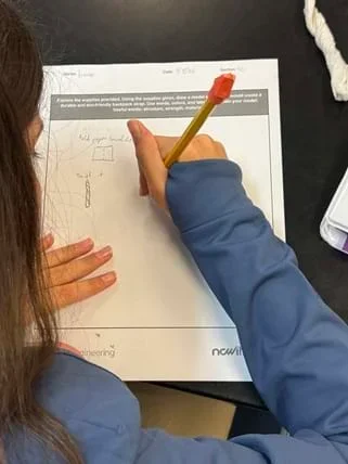 A photo taken over the shoulder of a person seated at a black tabletop, drawing on a printed worksheet with their right hand. The worksheet has printed text and small hand-drawn sketches. A crumpled paper towel is at the top right, and a white phone or case is partly visible at the right edge.
