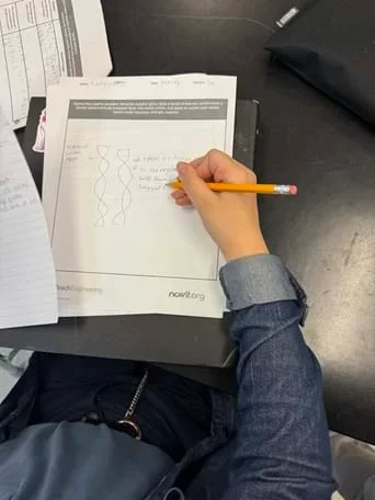 A photo taken over the shoulder of a person seated at a black table, using their right hand to draw on a printed worksheet. Loose papers and a black fabric pouch sit near the top-right edge of the photo.