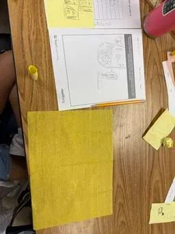 A photo showing the top of a classroom desk with activity planning sheet, as well as a pencil design on the left and a plain piece of yellow tissue paper on the right.