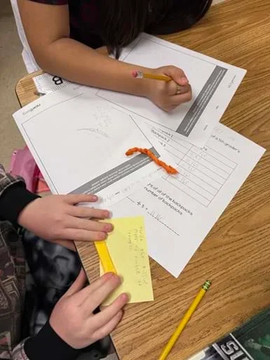 A photo of a student desk showing 2 sets of student hands working. One student is holding a pencil and writing on the planning worksheet, while the other student is folding tissue paper. There is another piece of tissue paper that has already been folded laying on the desk.