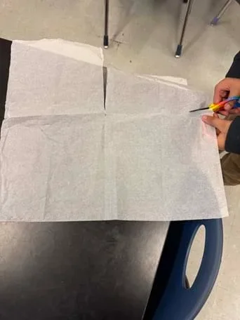 A photo showing an overhead view of a student cutting a large sheet of thin white tissue paper on a black tabletop. Two hands hold rainbow-handled scissors at the right edge of the sheet; a straight cut has been made near the top center. The scene suggests students building paper strap prototypes.