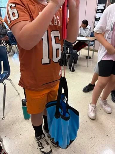 A photo showing a student holding a spring scale attached to a tissue paper prototype. The tissue paper is looped around a flexible bucket filled with weight to test the prototype.