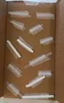 A photograph showing the inside of a shallow cardboard box. Several clear plastic straws are taped at angles along the bottom and sides of the box using pieces of clear tape. The straws are positioned randomly, some overlapping, with tape strips holding them in place. The box is empty aside from the taped straws.