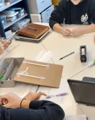 A photo showing a group of students working collaboratively at a white table. In the center, a piece of brown cardboard serves as a base for a structural prototype, with white plastic straws taped onto it in a geometric pattern. Surrounding the project are various supplies: a bundle of loose straws, a black tape dispenser, and a gray organizer bin containing scissors, a purple pen, and a laptop.