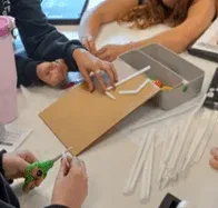 A photo showing students working collaboratively at a white table around a piece of brown cardboard. The cardboard has white plastic straws taped onto it. Surrounding the project are various supplies.