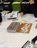 A photo showing students working collaboratively at a white table around a piece of brown cardboard. The cardboard has white plastic straws taped onto it. Surrounding the project are various supplies.