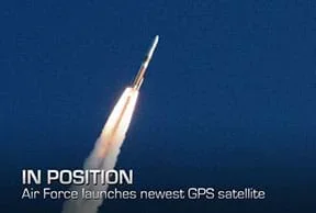 A photograph shows a blasting off rocket (equipped with a GPS satellite) as it propels through the air, trailing a stream of flame and smoke from the engines. The rocket is red on top and white on bottom with a red stripe in the middle. Overlaid text reads “IN POSITION. Air Force launches newest GPS satellite.”