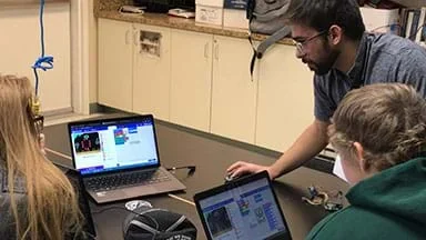 This image shows students sitting at a table with their laptop computers learning some basics of this lesson and its associated three-part activity.