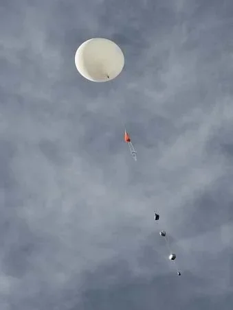 Looking up toward a dark blue sky with high white clouds at a white, round weather balloon that is carrying a long string of payload items below it, including a red parachute and four smaller boxes.