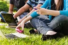 A photograph shows three teenagers sitting on the grass using laptops and cell phones and listening to music with ear buds.