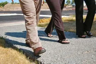 A photograph shows the legs and feet of three people walking on a gray sidewalk that looks like it is made of much aggregate, as spaces can be seen between the aggregate pieces.
