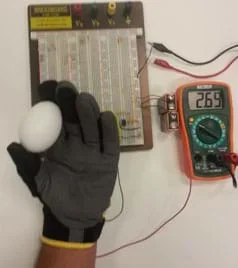 A photograph shows the same setup as Figure 4 but this time a hand is inside the glove and it holds a white egg between thumb and index finger. The connected multimeter reads 2.69.