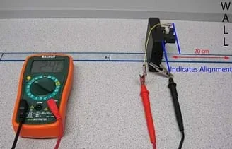 Photo shows a multimeter, measuring tape, and box to calibrate a Sharp GP2Y0A02YK0F Sensor. The sensor is facing the wall is connected to the multimeter by two wires, one red and one black with alligator clips.