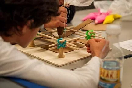 A photograph shows two students at a table focused on using a model/mock-3D bioprinter. One student squeezes an icing bag that represents the extrusion of cells, while the other student moves the cardboard printing stage in order to print an object of the desired shape.