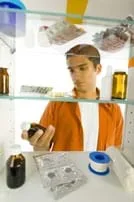 Photo shows young man looking into a medicine cabinet and holding a brown bottle of liquid medicine.