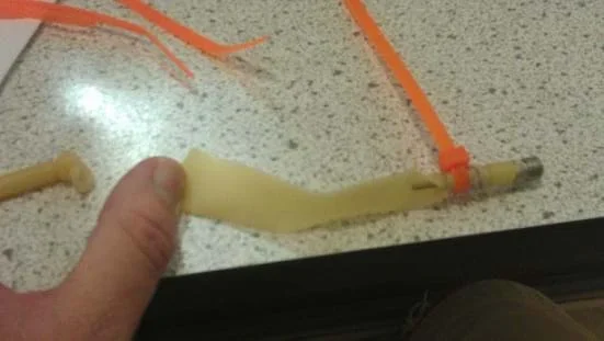 A photograph of a short piece of latex tubing attached to the needle tip extension of a bicycle pump with a zip tie and a rubber band. The latex tube is ripped from an explosion.