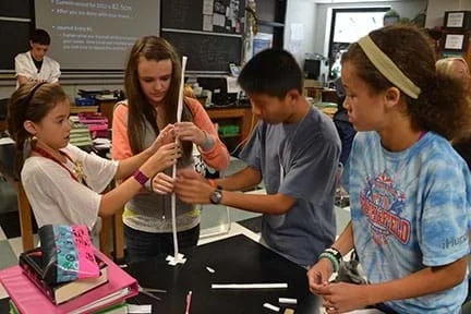 Four students are constructing a paper tower that is narrow, round, and about two feet tall.