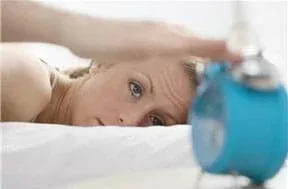 Photograph shows a woman waking up and reaching for an alarm clock near her bed.